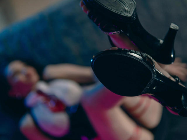 Le gambe di JennaDerose sono sollevate, indossa tacchi alti neri lucidi con punta, mostrando la suola, mentre è sdraiata sulla schiena, sfocata, con i suoi seni e glutei visibili sullo sfondo..