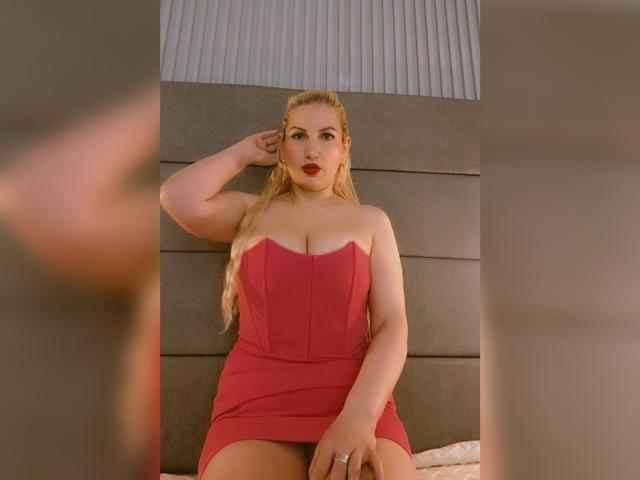 GiuliaAddams, avec des cheveux blonds, une robe rouge sans bretelles, de grandes seins, et une bague, la main dans les cheveux, assise, regardant la caméra, bras flous sur les côtés, éclairage doux, expression confiante..
