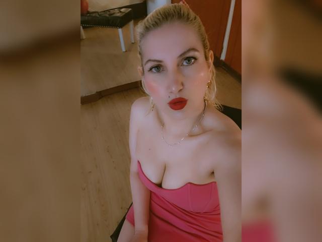 GiuliaAddams, avec des cheveux blonds, du rouge à lèvres et une robe rose sans bretelles, montrant ses seins, assise sur une chaise noire, avec un sol en bois et un miroir en arrière-plan..