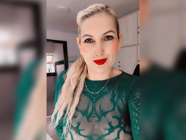 GiuliaAddams, avec des cheveux blonds attachés en queue de cheval, un haut en dentelle verte, du rouge à lèvres rouge, un anneau au nez et une chaîne en argent, souriant à la caméra. Ses seins sont visibles à travers la dentelle..