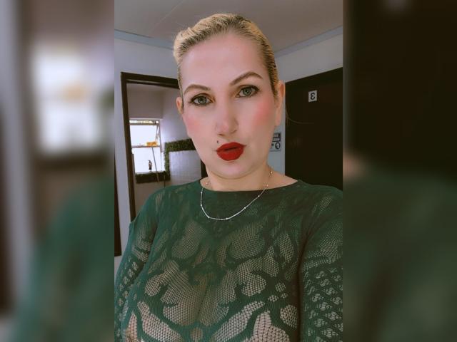 GiuliaAddams a les lèvres rouges, un haut en dentelle verte, et les cheveux blonds attachés en chignon. Elle regarde l'appareil photo avec ses yeux verts, et se tient dans une pièce avec des portes noires et blanches floues..
