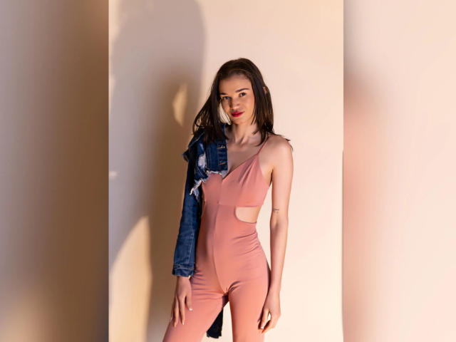 JennaDerose in a pink one-piece with cutouts, wearing a denim jacket on one arm, standing against a wall, red lipstick, long brown hair, slim, sexy, cute.