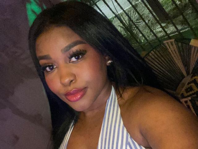 NicolZurich a la peau brun foncé, de longs cheveux noirs, et porte un haut à bretelles rayé. Ses lèvres sont d'un rose brillant, et elle a des sourcils épais et arqués. Elle sourit légèrement..