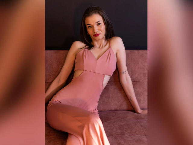 JennaDerose indossa un abito rosa con tagli e senza maniche, seduta su un divano rosa, con le gambe incrociate e le braccia appoggiate, rossetto rosso, capelli castani, tatuaggio sul braccio destro, sorridente..