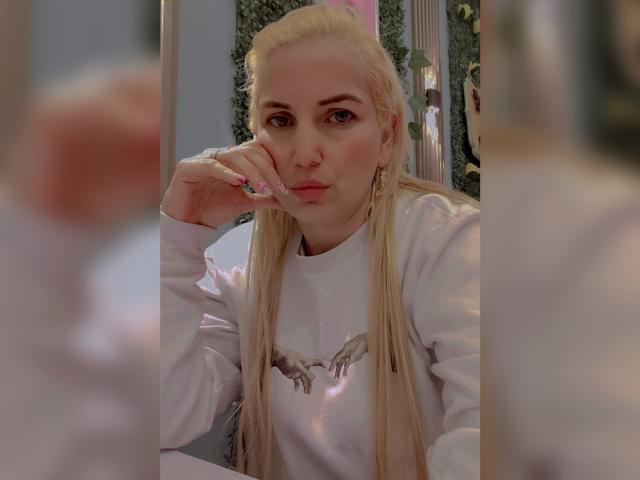 GiuliaAddams, blonde avec de longs cheveux, portant un sweat à capuche blanc, appuyant son menton sur sa main, regardant un miroir avec une légère moue, des ongles roses et des boucles d'oreilles..