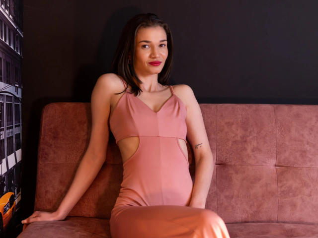 JennaDerose in un abito rosa con tagli, seduta su un divano rosa, che mostra le sue braccia sottili, il seno piccolo e il tatuaggio, con un leggero sorriso e rossetto rosso..