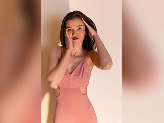 JennaDerose in a pink, cut-out dress, touching her face with hands, has red lipstick and brown hair, standing against a plain wall, with blurred reflections on both sides.