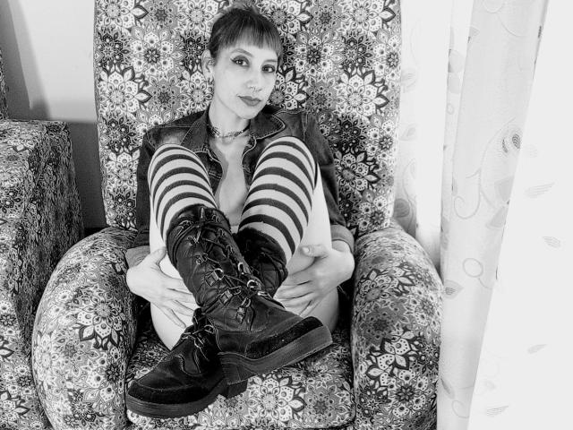 AliRousse sits in a floral armchair, legs up, wearing a leather jacket, striped thigh-high socks, and black lace-up boots, with a short, dark haircut and a serious expression.