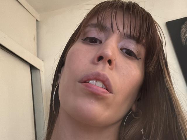 SeleneBright's face, showing a nose piercing, with brown hair, black eyeliner, and slightly parted lips, wearing large hoop earrings, looking up with a small, white-toothed grin.