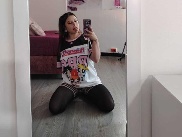 MelaniBecker, kneeling, wearing a loose white and black soccer shirt, black thigh-high stockings, taking a mirror selfie with a smartphone, tongue out, bedroom, wooden floor.