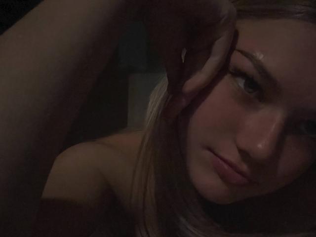 RandaDaill's face in a dimly lit photo, blonde hair, blue eyes, slightly parted lips, hand touching her forehead, bare shoulders, soft light on her skin, subtle expression.