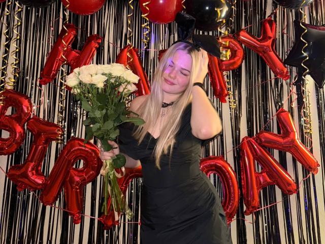 RandaDaill, with long blonde hair, wearing a black dress, holding white roses, standing in front of red balloons and black decorations, smiling with eyes closed.