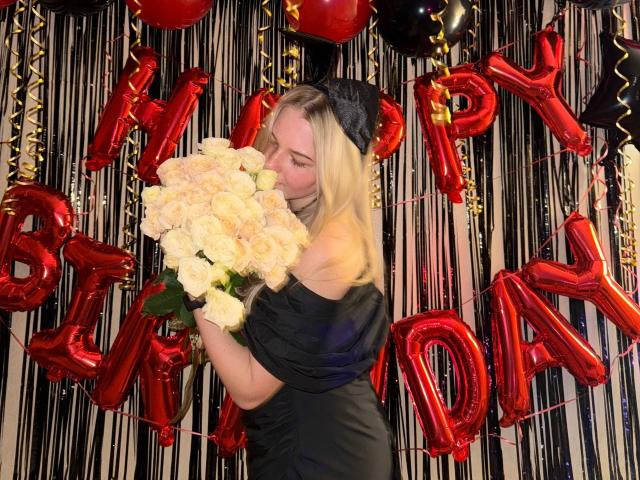 RandaDaill with blonde hair, black off-shoulder dress, black headband, kissing white roses, standing in front of red birthday balloons, black and white striped background, photo.
