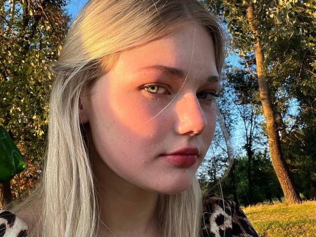 RandaDaill's face with pale skin, green eyes, and blonde hair in sunlight, wearing a leopard print shirt, has pink lips and a slightly serious expression.