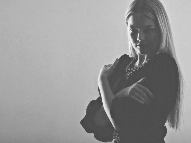 Black-and-white photo of RandaDaill with long blonde hair, arms crossed, wearing a dark top and necklace, looking down with a serious expression, showing her tits slightly.