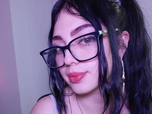 IsabellaaRossi with black glasses, red lipstick, and purple hair in a ponytail, wearing a choker and dangling earrings, smiling with slightly parted lips.