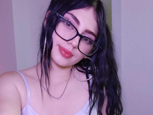 IsabellaaRossi with black hair, glasses, red lipstick, and gray tank top, wearing silver hoop earrings and necklace, smiling at the camera. Boobs visible.
