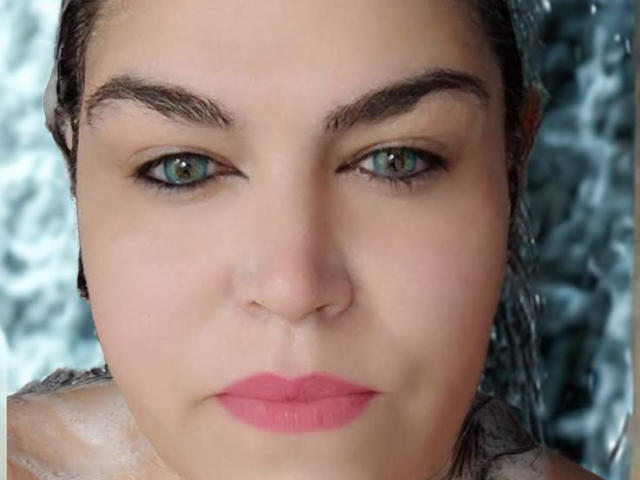 Het gezicht van NettyElvie, met groene ogen, roze lippenstift en nat bruin haar, die recht in de camera kijkt met een serieuze uitdrukking. Haar wenkbrauwen zijn donker en goed gedefinieerd..