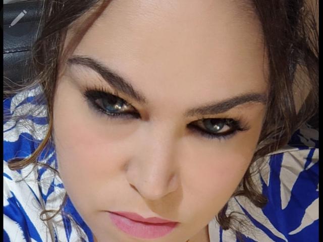 NettyElvie tem olhos azuis e expressivos, sobrancelhas escuras e grossas, e batom rosa. Ela está vestindo uma camisa com um padrão azul e branco, e tem cabelos castanhos cacheados. Seu nariz é ligeiramente arrebitado..