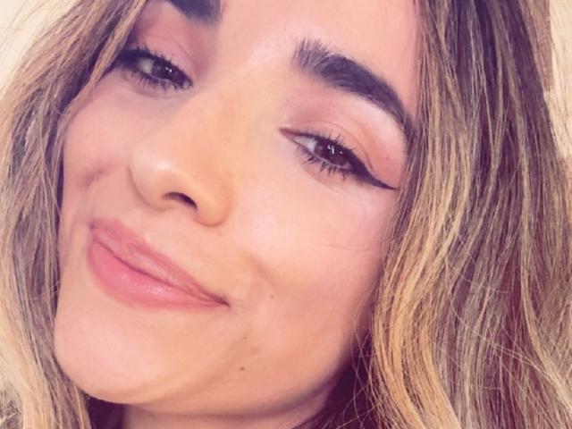 AdharaCooperr's face, with light brown hair, thick eyebrows, pink eyeshadow, winged eyeliner, and a subtle smile, showing a small acne mark below her right eye.