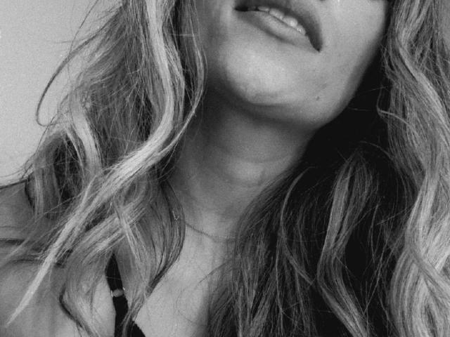 AdharaCooperr's face, showing her slightly parted lips with a hint of her teeth. Her wavy, blonde hair falls around her face. She's wearing a black top with thin straps.