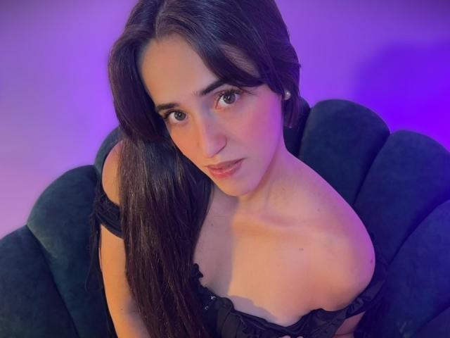 AlysonFox with long dark hair, looking up, wearing a black off-shoulder top, showing her small tits and bare shoulders, sitting on a green velvet chair.