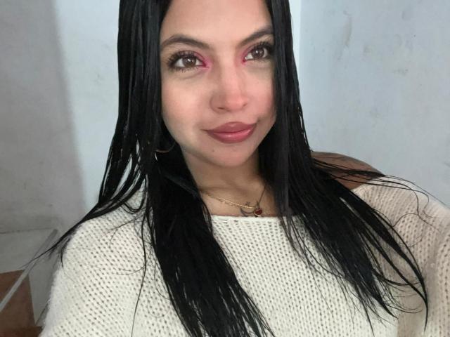 NyraVanel with black hair, pink eye makeup, and white sweater, smiling, has a small nose and is wearing a gold necklace. Boobs are not visible, only her upper body.