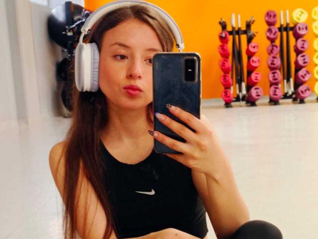 VioletaNova with long brown hair, wearing white headphones, black sports bra, and black leggings, pouting at her phone in a gym. She's taking a selfie with her phone.