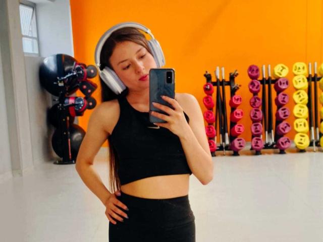 VioletaNova with small tits, wearing a black sports bra and black leggings, has her hand on her hip, taking a mirror selfie in a gym, with headphones on.