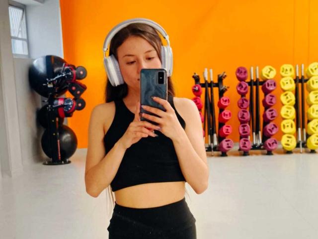 VioletaNova with small tits, wearing a black sports bra and headphones, taking a mirror selfie in a gym with colorful dumbbells in the background.