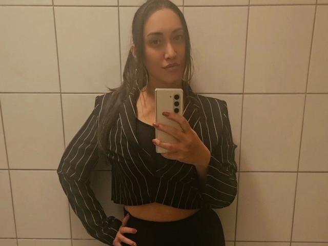 Bad-Bunnies, com cabelo longo e molhado, usando uma cropped preta com listras finas e calças pretas, tirando uma selfie no espelho com a mão na cintura, segurando um celular branco..