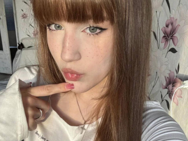 CorinaMakinster with pouty pink lips, finger on bottom lip, brown hair, green eyes, white shirt, red nails, and a fair skin tone, looking directly at the camera.