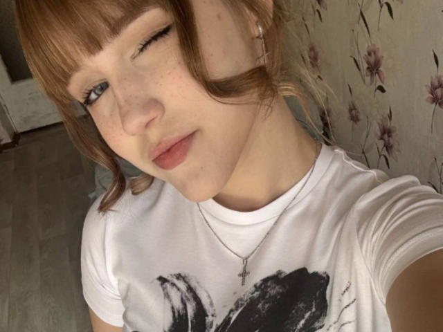 CorinaMakinster, winking, with light brown hair in pigtails, wearing a white t-shirt, cross necklace, and subtle makeup, showing freckles and soft pink lips, in a casual selfie.