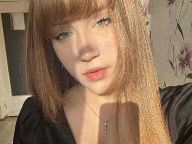 CorinaMakinster tiene el cabello castaño claro, liso y con flequillo, piel pálida, pecas, ojos verdes, labios rosados, y lleva una blusa negra con un collar de cruz, con la luz del sol en su rostro..