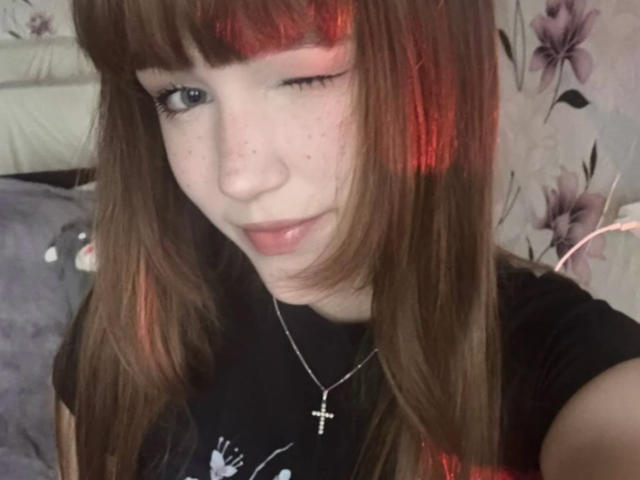 CorinaMakinster, con cabello castaño largo, reflejos rojos y flequillo, guiñando un ojo, vistiendo una camisa negra, un collar con una cruz y con pecas, sonriendo suavemente. Su trasero no es visible..