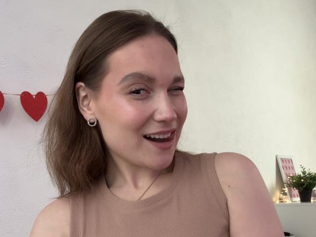 MarikoDentino with light brown hair, winking, smiling, wearing a beige sleeveless top, small hoop earrings, and a necklace, in a relaxed setting. Her tits are partially visible.