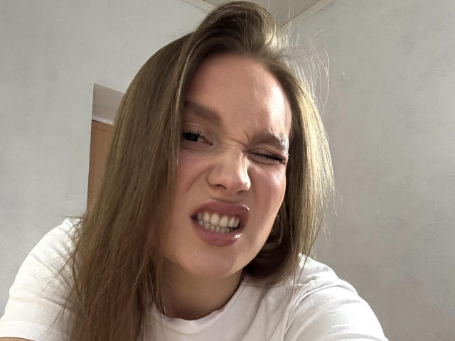 MarikoDentino with brown hair, winking one eye, and grimacing with clenched teeth, wearing a white shirt, looking slightly annoyed. Her nose is centered in the frame.