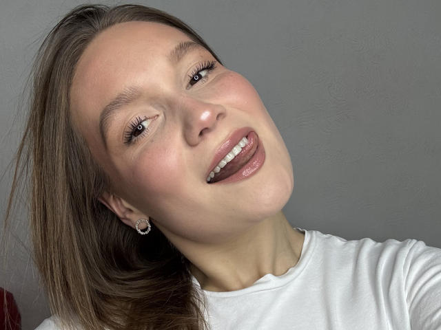 MarikoDentino has a cute face with long brown hair, wearing a white shirt, showing a happy smile with glossy pink lips, and cute earrings against a gray background.