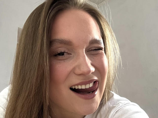 Mariko Dentino tiene el cabello castaño claro, y está guiñando, con la lengua ligeramente fuera, mostrando sus dientes blancos. Lleva una camisa blanca, y su rostro está sonrojado..