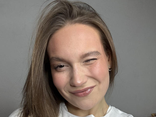 MarikoDentino tem um rosto adorável, com um sorriso malicioso, um olho fechado, lábios rosados e brilhantes e cabelo castanho. Ela está vestindo uma camisa branca e pequenos brincos..