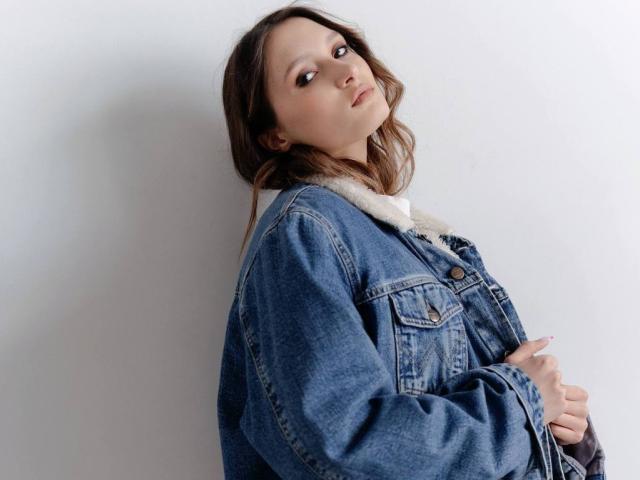 Maximina Condroski, com cabelos castanhos ondulados, vestindo uma jaqueta jeans azul com uma gola branca felpuda, olhando ligeiramente para o lado com uma expressão neutra, com as mãos perto do peito..