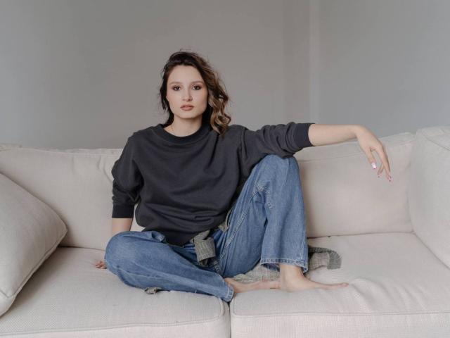 Maximina Condroski sitzt auf einem weißen Sofa, trägt einen grauen Pullover und blaue Jeans, ein Arm ruht am Rückenlehne, und ihre Beine sind gekreuzt, sie ist barfuß..