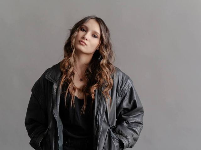 Maximina Condroski mit welligem, braunem Haar, trägt eine schwarze Lederjacke über einem schwarzen Oberteil, die Hände in den Taschen, blickt leicht zur Seite, grauer Hintergrund..