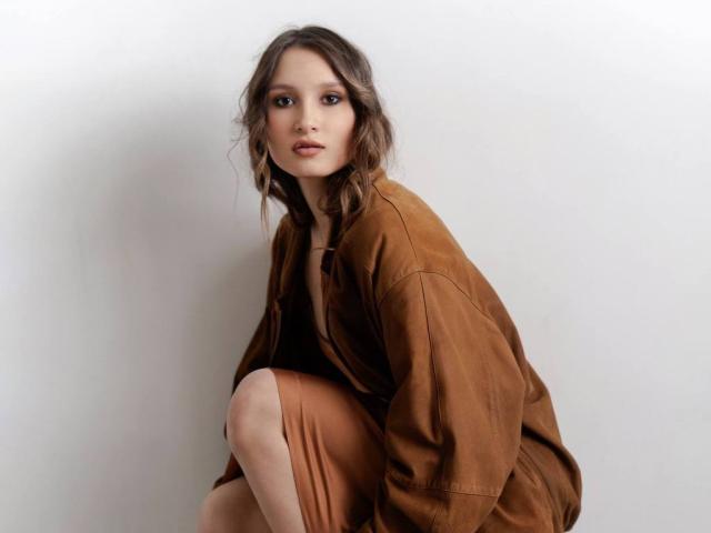 Maximina Condroski mit welligem, braunem Haar, trägt eine braune, übergroße Jacke und einen passenden Rock, sie hockt, blickt neutral in die Kamera, weiches Licht auf ihrem Gesicht..
