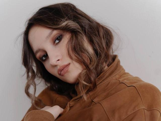 Maximina Condroski, met golvend bruin haar, verleidelijke ogen en een bruine suède jas, kijkt opzij met een lichtjes fronsende uitdrukking. Haar achterwerk is gedeeltelijk zichtbaar..