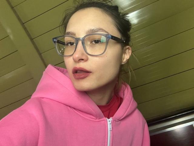 MaximinaCondroski with glasses, pink hoodie, and red shirt, showing slightly parted lips, brown hair tied back, and a neutral expression. Her ass is not visible.