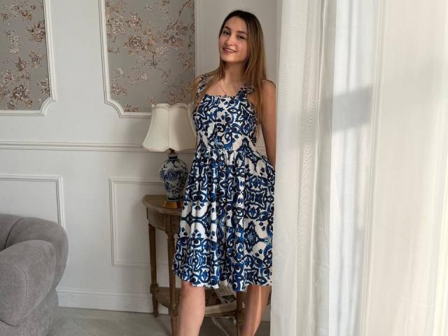 Maximina Condroski, con un seno di media grandezza, indossa un abito floreale blu e bianco, è in piedi accanto a una tenda bianca, sorride, ha i capelli castano chiaro, e si trova in una stanza con una luce soffusa..