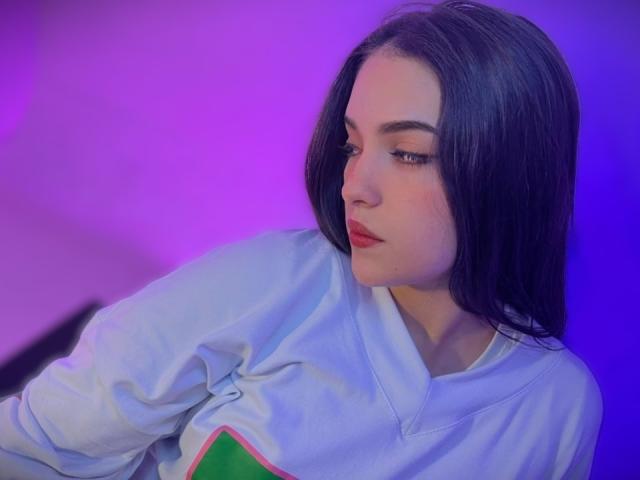 KatarinaNovak with long black hair, red lipstick, and a light blue shirt with green and pink accents, looking down with a slight smile, purple and blue lighting.