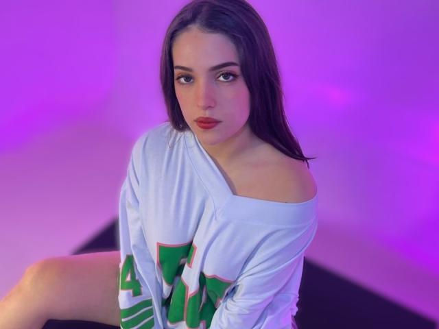 KatarinaNovak with long brown hair, off-shoulder white shirt, red lipstick, and green/white lettering, sitting with legs apart, looking at camera, purple lighting.