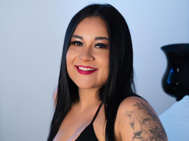 Chloe Rouse, com cabelos longos e pretos, sorrindo, usando batom vermelho, top de alça preta, mostrando tatuagem no ombro direito, piercing no nariz, olhos azuis, pele clara, olhando para a câmera..